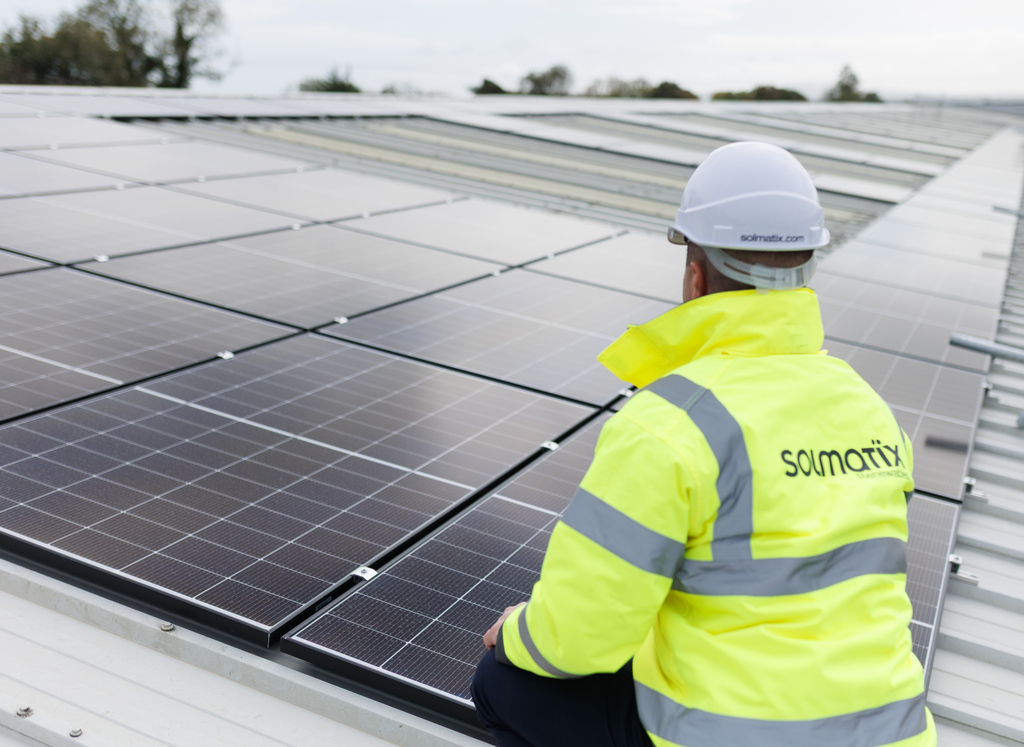 A solar technician wearing a white hard hat and high-vis jacket inspecting a large solar panel array on a commercial rooftop.