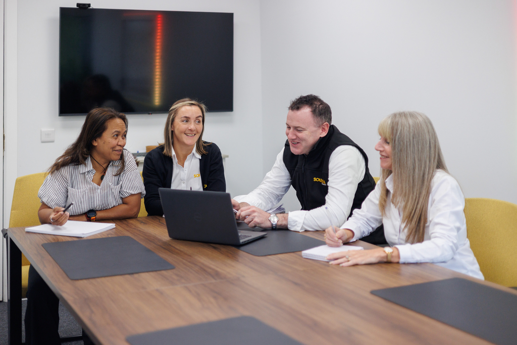 Three female colleagues and one male colleague sitting together at a table discussing paperwork and looking at a laptop computer.
