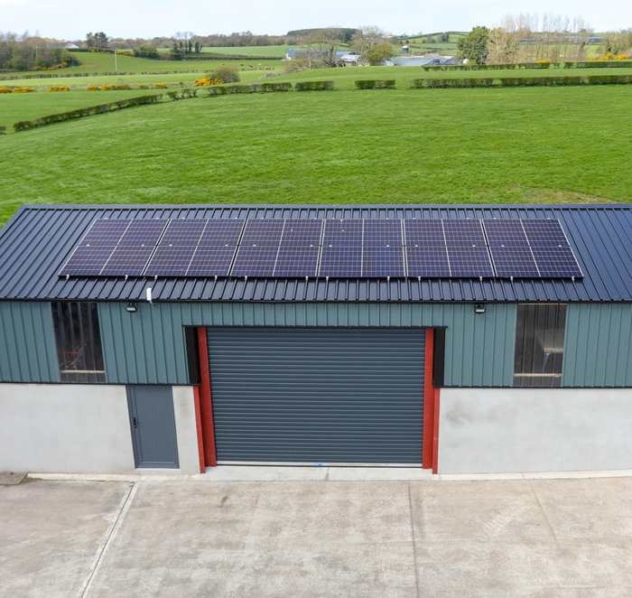 An aerial shot of solar panels mounted on the dark metal roof of a rural agricultural shed overlooking green fields.