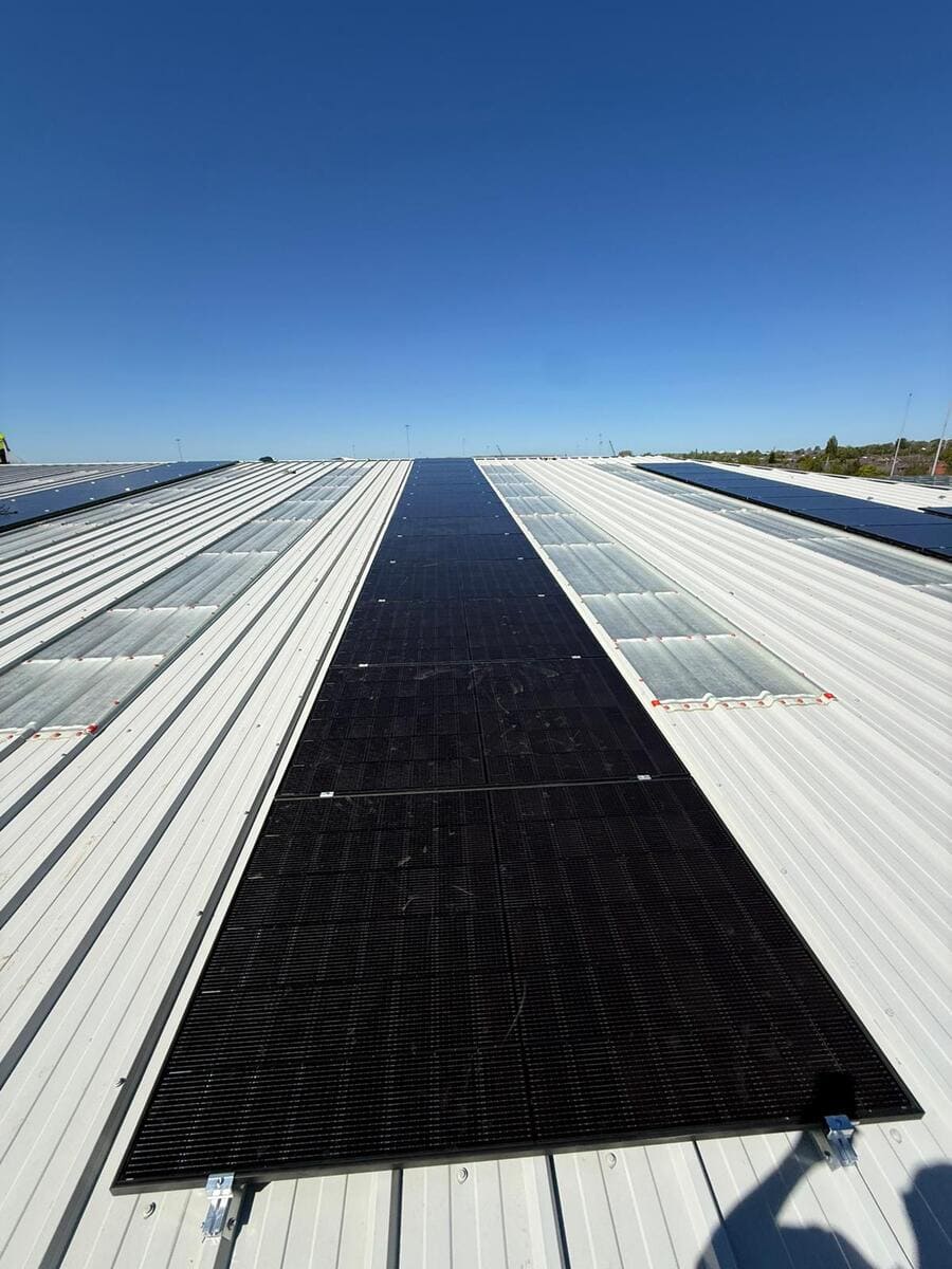Wide prospective view looking down a long strip of solar panels installed on a white metal roof under a bright blue sky.