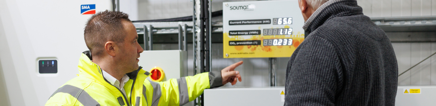 Technician pointing toward a wall-mounted solar monitoring display while speaking with a client.